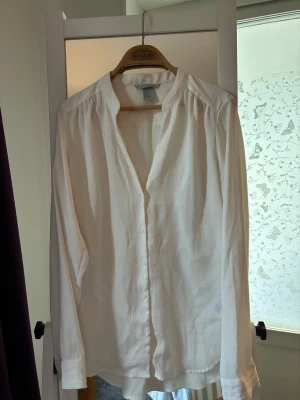 Vit klassisk långärmad blus - Stilren vit blus med knappar framtill och lätt rynkade axlar. Blusen har en rak passform, enkel krage och långa ärmar med manschett. Perfekt att matcha till jeans eller kjol för en clean look.