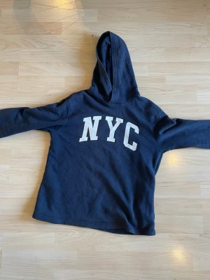Mörkblå hoodie med NYC-tryck - Snygg mörkblå hoodie med stor vit NYC-text på framsidan. Klassisk modell med huva och långa ärmar. Perfekt för dig som gillar streetwear och vill ha en enkel men stilren look. Mjukt material. Storlek 11-12 år 146/152. Material: bomull, polyester 