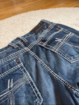 Blå jeans med kontrastsömmar - Säljer ett par blå jeans med tydliga vita kontrastsömmar och coola broderade detaljer på bakfickorna. Lite kortare LÄNGD en vad som står!