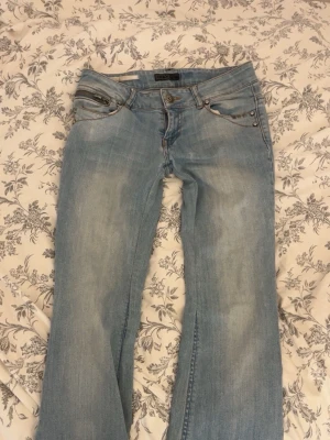 Ljusblå bootcut jeans med nitar - Säljer ett par ljusblå bootcut jeans med slitningar och snygga detaljer på bakfickorna. Fickorna har dekorativa sömmar och silverfärgade nitar. Jeansen har klassisk femficksmodell och är tillverkade i denim med lätt stretch.
