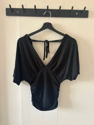 Svart topp gina v ringning stl s - Snygg svart topp med djup v-ringning och knyt i ryggen. Toppen har korta, vida ärmar och smickrande passform som framhäver midjan. Perfekt att styla med både jeans och kjol för en trendig look. Använd en gång, skriv vid funderingar 💞