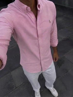 Rosa skjorta från Polo Ralph Lauren - Säljer en klassisk rosa skjorta från Polo Ralph Lauren i storlek M. Skjortan har button-down krage, långa ärmar och den ikoniska broderade loggan på bröstet. Tillverkad i ett mjukt bomullstyg som känns riktigt skönt mot huden. Skjortan har inga defekter och pris kan diskuteras.