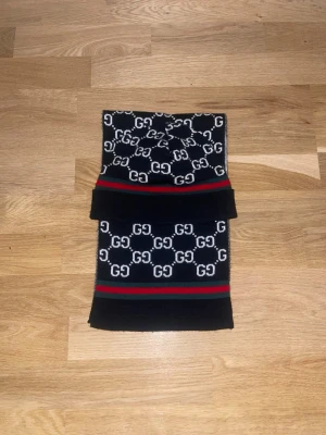 Svart och vit halsduk med GG-mönster - Snygg halsduk i svart och vitt med klassiskt GG-mönster och ränder i rött och grönt. Halsduken har en rektangulär form och är stickad i mjukt material. Perfekt för dig som vill ha en trendig accessoar med lyxig känsla.