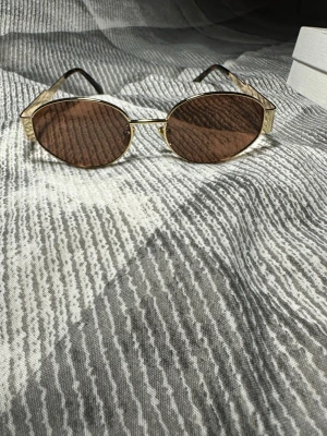 Guldiga ovala solglasögon från Celine - Snygga solglasögon från Celine med guldfärgade metallbågar och bruna ovala glas. Bågarna har en unik struktur och Celine-loggan på sidorna. Perfekta för dig som vill ha en lyxig och trendig accessoar till sommaren.