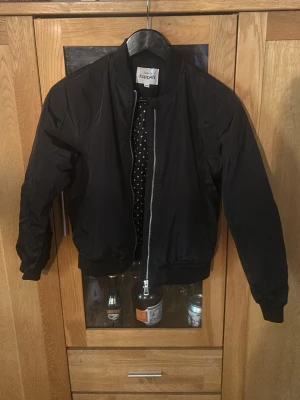 Svart bomberjacka från KappAhl - Snygg svart bomberjacka från KappAhl med silvrig dragkedja och ribbade muddar. Jackan har ett coolt prickigt innerfoder som ger extra stilpoäng. Perfekt för dig som vill ha en klassisk men trendig look.