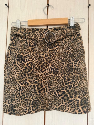 Leopardmönstrad kortkjol med bälte - Snygg kortkjol med leopardmönster och tillhörande bälte med rund metallspänne. Kjolen har en beige och svart färgskala och är tillverkad i ett mjukt polyestertyg.