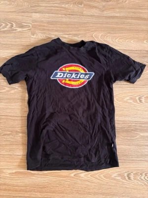 Svart Dickies t-shirt med logga - Säljer en svart t-shirt från Dickies med den klassiska färgglada loggan tryckt på bröstet. T-shirten har rund hals och korta ärmar. Perfekt till dig som gillar streetwear och enkel stil.