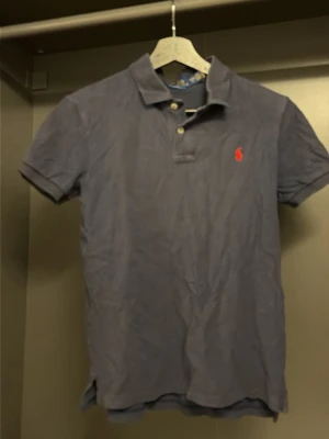 Marinblå pikétröja från Polo Ralph Lauren - Klassisk marinblå pikétröja från Polo Ralph Lauren i slim fit-modell. Tröjan har korta ärmar, krage med knappar och den ikoniska röda logotypen broderad på bröstet. Tillverkad i bomull för en bekväm känsla.