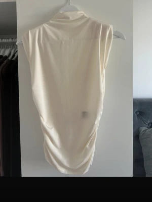 Krämvit ärmlös polotopp - Säljer en krämvit ärmlös polotopp med mjukt, stretchigt material och draperad effekt längs sidorna. Toppen har en hög krage och en clean, minimalistisk look som passar till många olika stilar. Axelvaddar och är använd 2-3 gånger i storlek xs