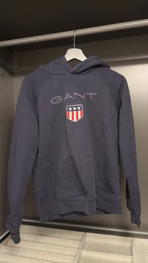 Mörkblå hoodie från GANT - Snygg mörkblå hoodie från GANT med klassisk logga och amerikanskinspirerat sköldtryck på bröstet. Hoodien har huva, magficka och ribbade muddar vid ärmar och nederkant. Perfekt för en avslappnad och sportig stil.