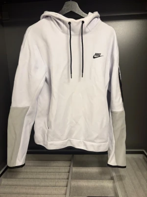 Vit och grå hoodie från Nike - Snygg vit hoodie från Nike med gråa partier på underarmarna och svarta dragsnören i huvan. Klassisk Nike-logga på bröstet. Hoodien har en avslappnad passform och är tillverkad i mjukt material, perfekt för en sportig och casual stil. Har en pytte liten fläck som går bort med lite noggrann tvätt 