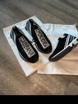 Svarta sneakers Dolce & Gabbana - Snygga svarta sneakers från Dolce & Gabbana med glittrande stenar och tydliga logotexter på ovansidan. Skorna har vita och svarta detaljer, elastisk slip-on design och en chunky sula för extra attityd.