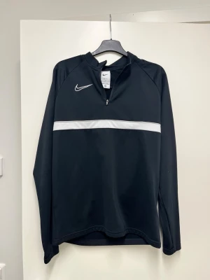 Svart Nike träningströja half zip 170 - Snygg svart långärmad träningströja från Nike med vit rand över bröstet och klassisk Nike-logga. Tröjan har half zip och är tillverkad i funktionsmaterial som andas, perfekt för träning eller uppvärmning.