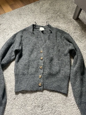 Mörkgrå kofta från H&M med hjärtknappar - Mysig mörkgrå stickad kofta från H&M med långa ärmar och V-ringning. 