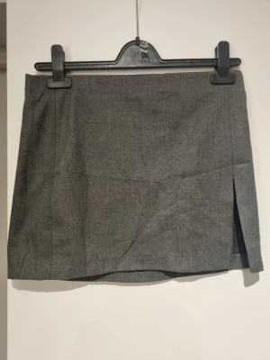 Grå kortkjol från Bershka - Stilren grå kortkjol från Bershka med en enkel design och diskret slits framtill. Kjolen har en rak passform och är tillverkad i ett mjukt material som känns bekvämt. Perfekt att matcha med en stickad tröja eller t-shirt.