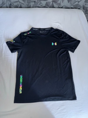 Svart t-shirt från Under Armour - Svart t-shirt från Under Armour med färgglada loggor och textdetaljer på bröstet, ärmen och längs sidan. T-shirten har rund halsringning och är tillverkad i ett lätt och sportigt material, perfekt för en aktiv livsstil.
