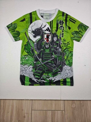 Grön samuraj t-shirt med drakmotiv - Unik grön t-shirt med svart och vitt tryck av samurajer och en drake på både fram- och baksida. T-shirten har korta ärmar, vita muddar och coola japanska detaljer. Perfekt för dig som gillar streetwear och vill sticka ut med ett färgstarkt motiv.
