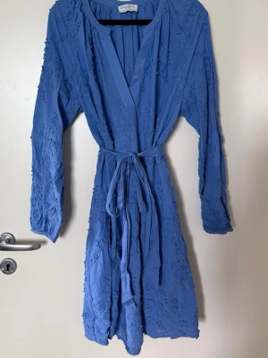 Blå omlottklänning med broderi - Snygg blå omlottklänning med långa ärmar och knytskärp i midjan. Klänningen har fina broderade blommor över hela tyget och en djup v-ringning. Perfekt för dig som gillar en avslappnad men ändå trendig stil.
