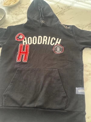 Hoodrich  - Svart hoodie från Hoodrich med vit broderad logga och röda detaljer. Stora tryck och patches framtill, känguruficka och ribbade muddar. Klassisk huva med dragsko och mjukt material som känns skönt mot huden.