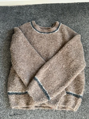 Brun fluffig stickad tröja - Mysig beige stickad tröja med fluffig textur och rund hals. Tröjan har mörkgrå och vita ränder vid hals, ärmslut och nederkant. Perfekt för kalla dagar och enkel att matcha med jeans eller kjol.