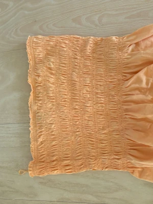 Ljus orange bandeau tubtopp Gina Tricot - Ljus orange bandeau tubtopp från Gina Tricot i smockat tyg. Toppen är ärmlös och croppad med en mjuk, stretchig känsla. Perfekt för sommardagar och enkel att matcha med olika stilar.