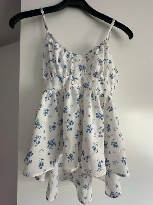 Vit blommig linnetopp med knyt - Supersöt vit linnetopp med blå blommor och tunna axelband. Toppen har en flowy passform och knyt i ryggen som ger en extra detalj. Använd 3 gånger. 