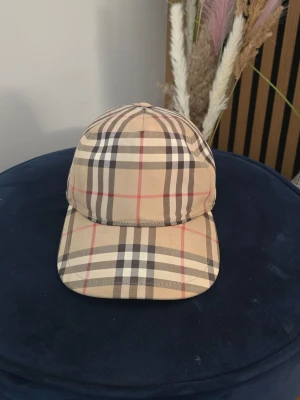 Beige keps från Burberry med rutmönster - Snygg beige keps från Burberry med det klassiska rutmönstret i svart, vitt och rött. Justerbar baktill med svart spänne och tydlig Burberry London England-logga. Perfekt accessoar för dig som gillar stilrena och tidlösa detaljer.