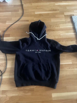 Svart hoodie från Tommy Hilfiger - Snygg svart hoodie från Tommy Hilfiger med vit dragsko i huvan och klassisk logga samt 'EST. 1985' tryckt på bröstet. Stor känguruficka framtill och ribbade muddar. Perfekt för en chill och stilren look.