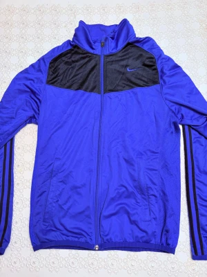 Blå Nike träningsjacka  - Snygg blå träningsjacka från Nike med svarta meshpaneler över bröstet och diskret Nike-logga. Jackan har hel dragkedja, hög krage och två sidofickor. Ränder längs ärmarna ger en sportig look. Perfekt för träning eller chill.