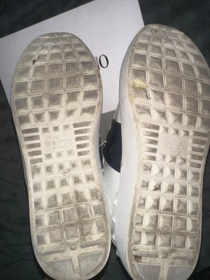 Valentino vita sneakers med svart detalj - Snygga vita sneakers från Valentino med svart detalj på sidan. Skorna har en klassisk låg siluett och är tillverkade i läder med gummisula. Perfekta för dig som vill ha en clean och exklusiv look.