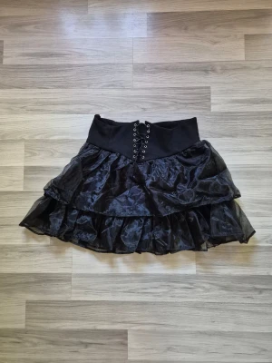 Svart volangkjol med snörning - Säljer en svart kortkjol med bred midja och snörning framtill. Kjolen har flera lager volanger i ett glansigt, tunt material som ger en cool och edgy look. Perfekt för dig som gillar alternativ stil och vill sticka ut.