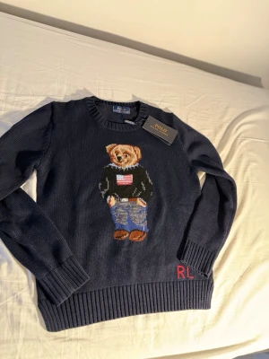 Ralph Lauren Mörkblå stickad tröja Polo Bear - Mörkblå stickad tröja från Polo Ralph Lauren med den ikoniska Polo Bear på bröstet. Tröjan har rund hals, ribbstickade muddar och RL-broderi nertill. Perfekt för dig som gillar klassisk och lekfull stil.