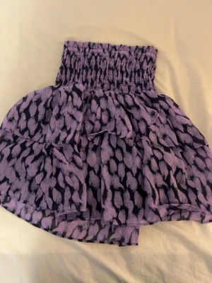 Lila kortkjol med volanger - Säljer en lila kortkjol med svart mönster och bred smock i midjan. Kjolen har flera lager volanger och ett unikt abstrakt mönster som ger en cool look. Perfekt för dig som vill sticka ut lite extra.