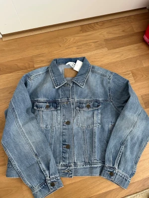 Blå jeansjacka från H&M - Klassisk blå jeansjacka från H&M med raka sömmar, två bröstfickor med lock och knappar samt knappstängning framtill. Jackan har en tidlös design och är tillverkad i slitstarkt denimtyg. Perfekt att slänga över en hoodie eller t-shirt.
