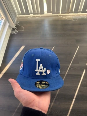 Blå LA Dodgers keps med brodyr - Säljer en blå snapback-keps med LA Dodgers-logga i vitt framtill och ett litet hjärta bredvid. På sidan finns en broderad patch med '1988 World Series'. Kepsen har platt skärm och klassisk passform. 