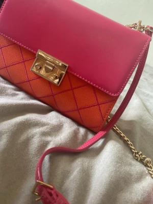 Rosa och orange quiltad handväska - Säljer en snygg handväska i rosa och orange med quiltat mönster och guldfärgat lås. Väskan har en axelrem i kedja och läder, samt en liten ficka på baksidan. Perfekt för dig som gillar färg och trendig design.