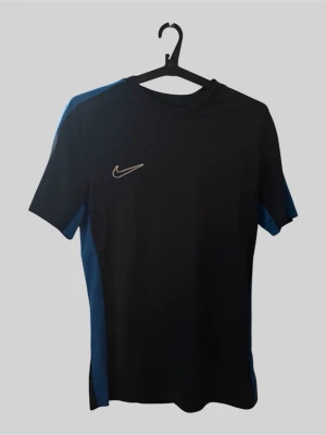 Svart och blå Nike träningströja - Snygg svart träningströja från Nike med blå detaljer längs sidorna och axlarna. Tröjan har korta ärmar, rund halsringning och är tillverkad i ett lätt och funktionellt material som passar perfekt för träning eller sport.