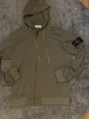 Olivgrön hoodie från Stone Island - Säljer en olivgrön hoodie från Stone Island med dragkedja framtill, två stora fickor och klassisk patch på ärmen. Tröjan har huva med dragsko och ribbade muddar. Perfekt för dig som gillar streetwear och vill ha något stilrent. M på pappret passar som L