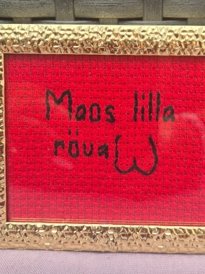 Tavla med busigt broderi  - En humoristisk broderad tavla med texten 'Maos lilla röva' på röd bakgrund. Tavlan har en dekorativ guldfärgad ram med struktur. Perfekt som rolig inredningsdetalj eller present.