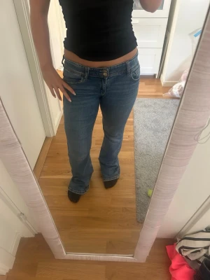Blå bootcut jeans med fickdetaljer - Säljer ett par klassiska blå bootcut jeans med låg midja och snygga fickdetaljer bak. Jeansen har en lätt utsvängd passform och är perfekta för en avslappnad men trendig look. Tillverkade i denim med kontrastsömmar och knappstängning framtill.
