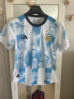 Argentina VM-tröja blåvit Adidas - Säljer en blåvit Argentina fotbollströja från Adidas med tryck av spelare och firande motiv på framsidan. Tröjan har officiella AFA-emblemet, svarta Adidas-loggan och texten 'CAMPEÓN MUNDIAL' samt spelarlista på ryggen. Klassisk rund hals och korta ärmar.
