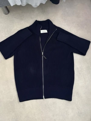 Mörkblå stickad ziptröja Maison Margiela - Snygg mörkblå stickad tröja från Maison Margiela med korta ärmar och silverfärgad dragkedja framtill. Tröjan har ribbad struktur och hög krage för en modern look. Perfekt till lager på lager och enkel att styla till olika outfits. Priset kan diskuteras 🍾