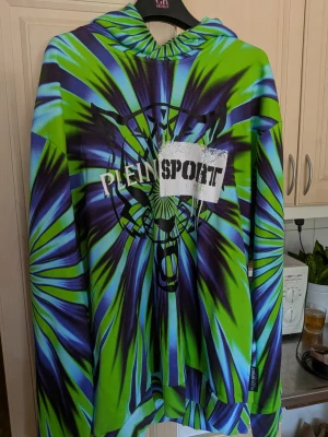 Färgglad hoodie från Plein Sport - Säljer en riktigt cool hoodie från Plein Sport med grön, blå och svart tie-dye mönster. Stor logga och tigertryck på bröstet. Hoodien har huva och långa ärmar, perfekt för dig som vill sticka ut med din stil. Storlek 4XL. Liten i storlek. 