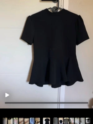 Svart peplum topp med kort ärm - Säljer en svart peplum topp med korta ärmar och rund hals. Toppen har en markerad midja och volang nedtill som ger en snygg siluett. Perfekt att styla med jeans eller kjol för en trendig look.