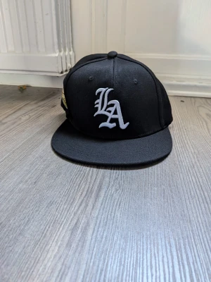 Svart LA snapback keps - Snygg svart snapback keps med stor vit LA-logga broderad framtill. Klassisk platt skärm och justerbar baksida.  Perfekt för dig som gillar streetwear och vill ha en stilren accessoar till din outfit.