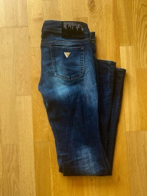 Mörkblå vintage jeans från Guess - Snygga mörkblå jeans från Guess med klassisk femficksmodell och diskreta slitningar framtill. Jeansen har raka ben och en liten metallogo på bakfickan. Materialet är stretchigt denim som sitter bekvämt och följer kroppen.