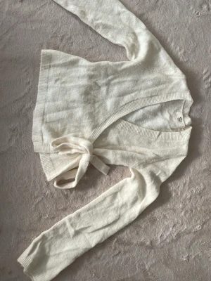 Vit omlottkofta med knytband - Säljer en mjuk och stilren vit stickad omlottkofta med långa ärmar och breda ribbade muddar. Koftan har ett knytband i midjan för en snygg passform och är perfekt för lager på lager. Passar till många olika outfits.