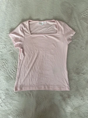 Ljusrosa topp med fyrkantig ringning - Söt ljusrosa topp med korta ärmar och trendig fyrkantig ringning. Tillverkad i ett mjukt, stretchigt material som sitter skönt mot kroppen. Enkel och stilren design som passar perfekt till jeans eller kjol. Använt 3 gånger men är nytvättad.