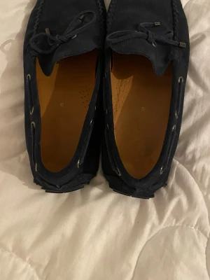 Mörkblå mocka loafers med snörning - Säljer ett par mörkblå loafers i mocka med dekorativ snörning framtill och metalländar på snörena. Skorna har klassisk seglarsko-design med detaljer längs sidorna och en mjuk innersula i brunt. Perfekta för en stilren och avslappnad look.