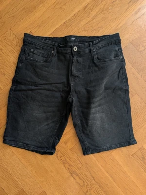 Svarta jeansshorts från Smog - Säljer ett par svarta jeansshorts från Smog med klassisk femficksdesign och knappgylf. Shortsen har en rak passform och är tillverkade i ett slitstarkt denimtyg. Perfekta för dig som gillar en enkel och stilren look.